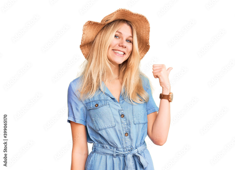 Young beautiful blonde woman wearing summer hat and dress smiling with happy face looking and pointing to the side with thumb up. Young beautiful blonde woman wearing summer hat and dress smiling with happy face looking and pointing to the side with thumb up.