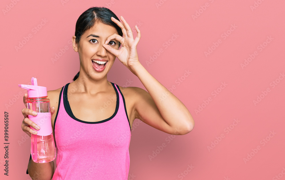 Beautiful asian young sport woman wearing sportswear drinking bottle of water smiling happy doing ok sign with hand on eye looking through fingers