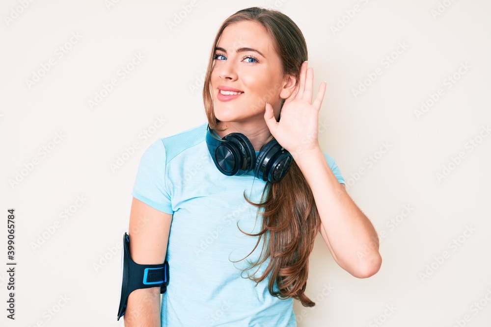 Beautiful young caucasian woman wearing gym clothes and using headphones smiling with hand over ear listening an hearing to rumor or gossip. deafness concept. Beautiful young caucasian woman wearing gym clothes and using headphones smiling with hand over ear listening an hearing to rumor or gossip. deafness concept.