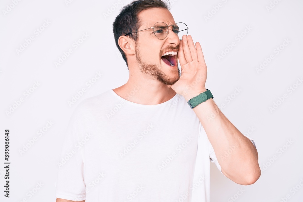 Young handsome man wearing casual clothes and glasses shouting and screaming loud to side with hand on mouth. communication concept. Young handsome man wearing casual clothes and glasses shouting and screaming loud to side with hand on mouth. communication concept.
