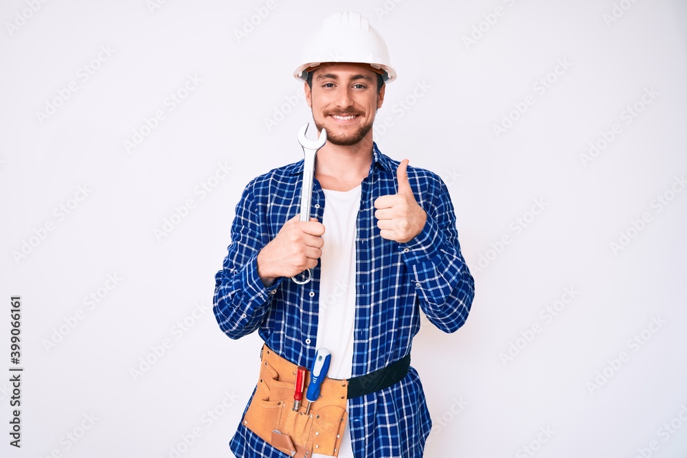 Young handsome man weaing handyman uniform holding wrench smiling happy and positive, thumb up doing excellent and approval sign