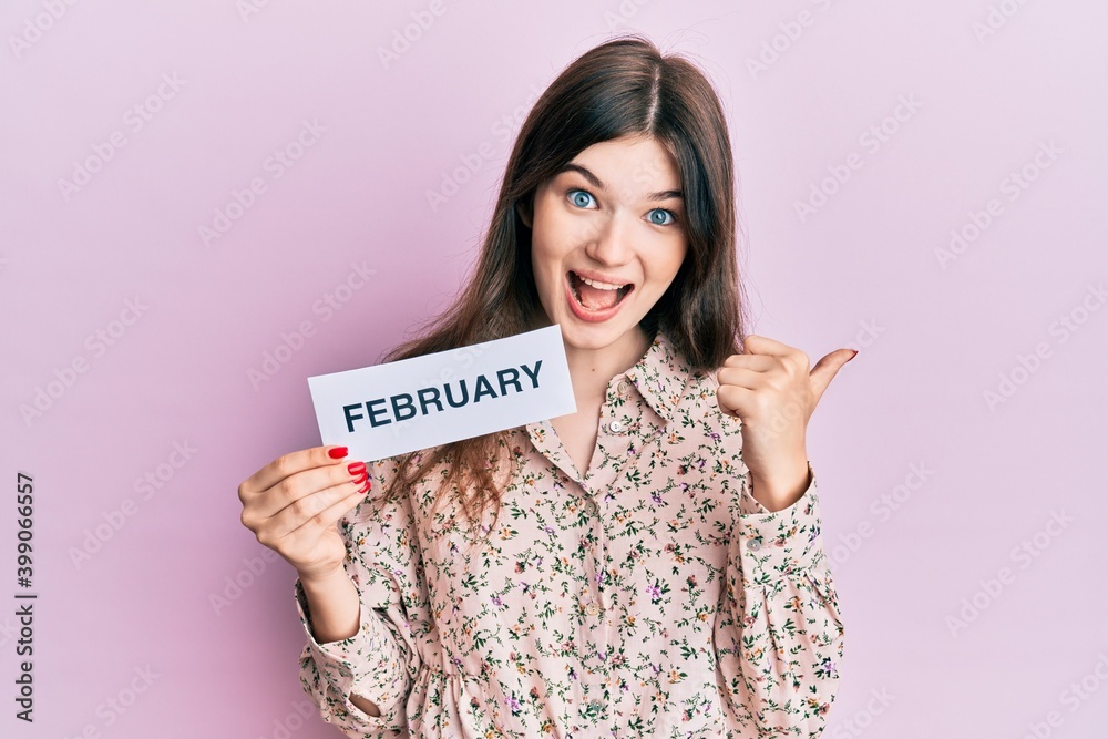 Young beautiful caucasian girl holding february word paper pointing thumb up to the side smiling happy with open mouth Young beautiful caucasian girl holding february word paper pointing thumb up to the side smiling happy with open mouth