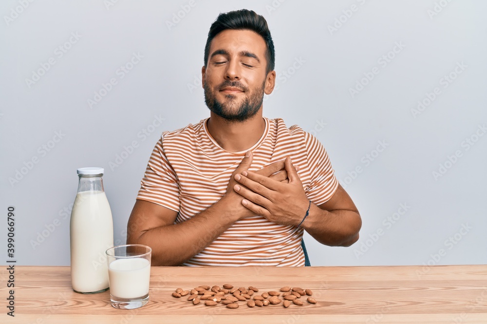 Handsome hispanic man drinking healthy almond milk smiling with hands on chest, eyes closed with grateful gesture on face. health concept. Handsome hispanic man drinking healthy almond milk smiling with hands on chest, eyes closed with grateful gesture on face. health concept.