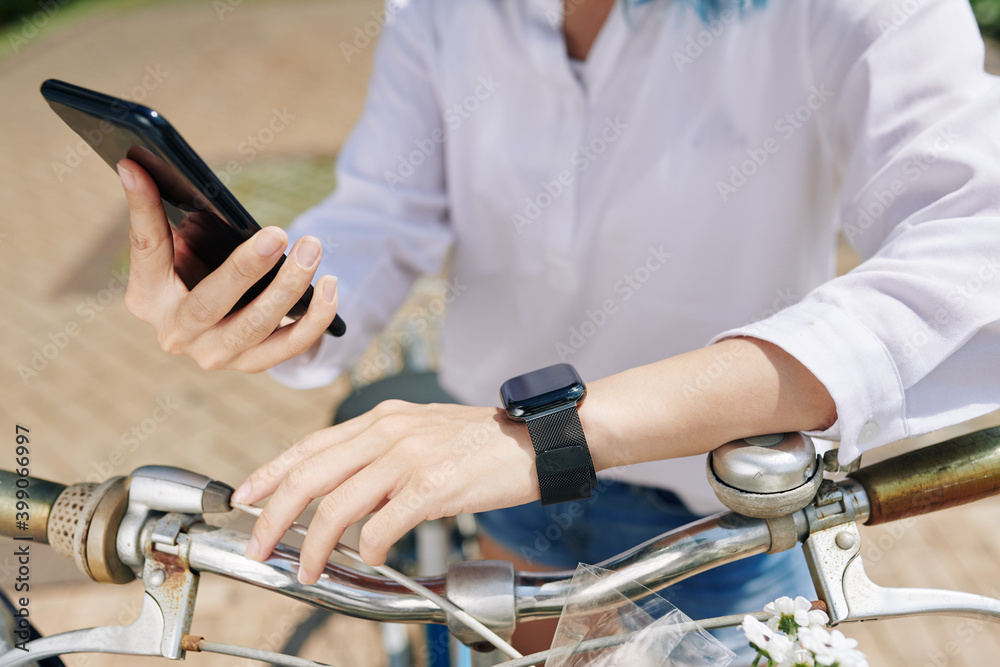 Close-up image of woman with smartwatch sitting on bicycle and checking applications on smartphone and smartwatch