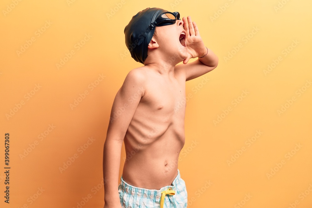 Cute blond kid wearing swimwear and swimmer glasses shouting and screaming loud to side with hand on mouth. communication concept.