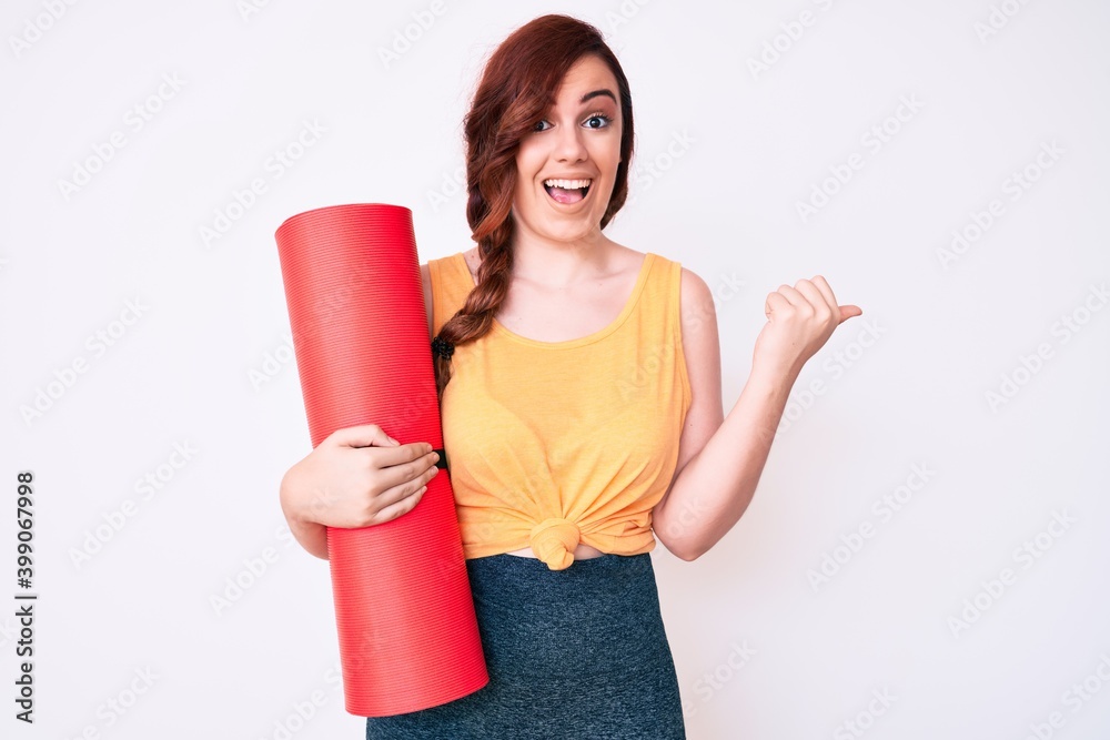 Young beautiful woman holding yoga mat pointing thumb up to the side smiling happy with open mouth Young beautiful woman holding yoga mat pointing thumb up to the side smiling happy with open mouth