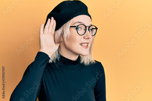 Young blonde girl wearing french look with beret smiling with hand over ear listening an hearing to rumor or gossip. deafness concept.
