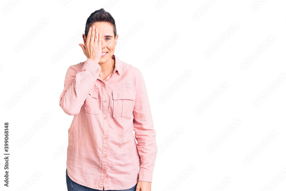 Young woman with short hair wearing casual clothes covering one eye with hand, confident smile on face and surprise emotion. Young woman with short hair wearing casual clothes covering one eye with hand, confident smile on face and surprise emotion.