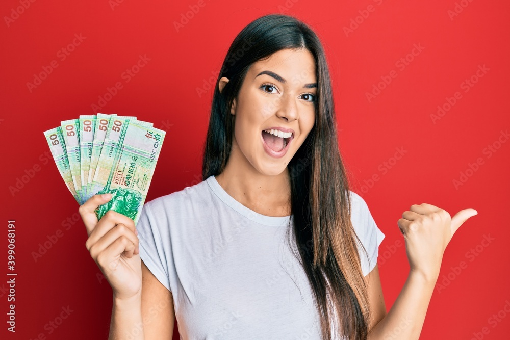 Young brunette woman holding hong kong 50 dollars banknotes pointing thumb up to the side smiling happy with open mouth Young brunette woman holding hong kong 50 dollars banknotes pointing thumb up to the side smiling happy with open mouth