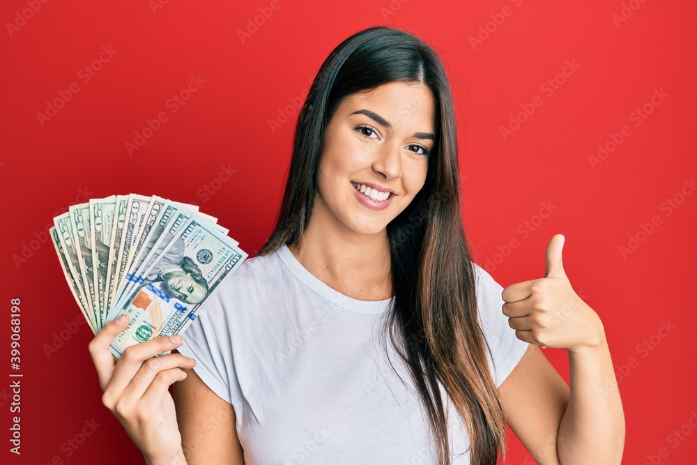 Young brunette woman holding dollars smiling happy and positive, thumb up doing excellent and approval sign Young brunette woman holding dollars smiling happy and positive, thumb up doing excellent and approval sign