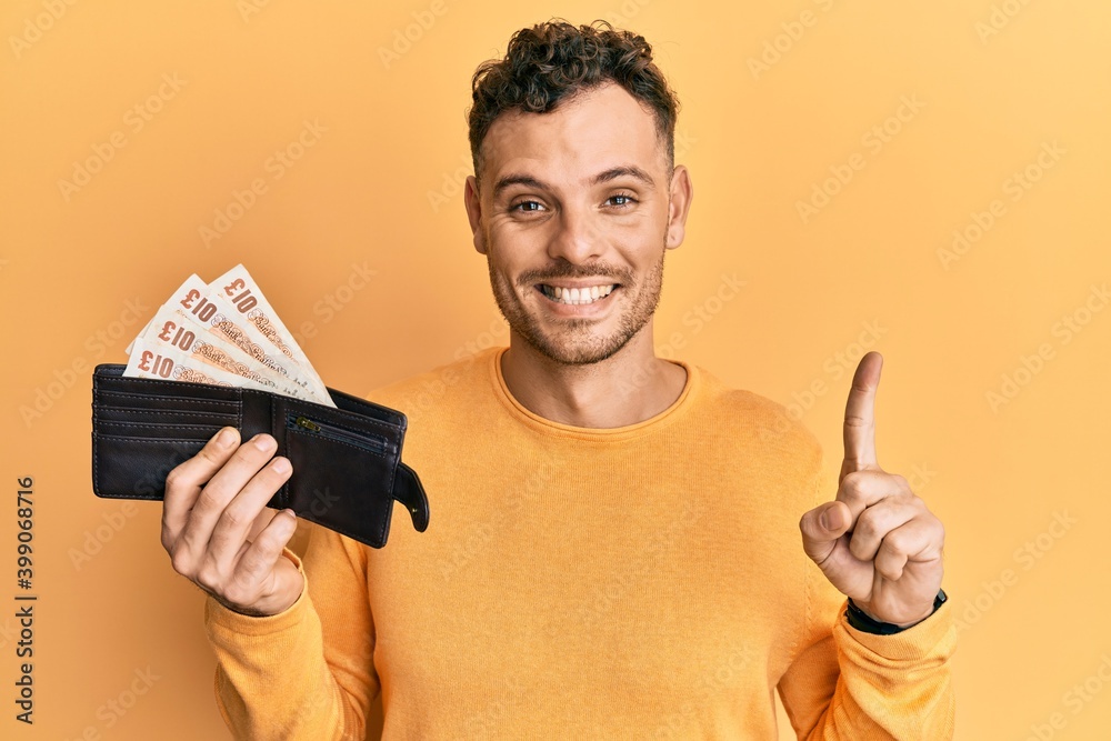 Young hispanic man holding wallet with united kingdom pounds smiling with an idea or question pointing finger with happy face, number one Young hispanic man holding wallet with united kingdom pounds smiling with an idea or question pointing finger with happy face, number one