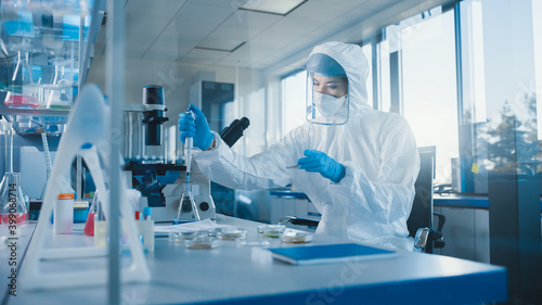 Beautiful Female Medical Scientist Wearing Coverall and Face Mask Using Micro Pipette while Working with Petri Dish. Vaccine  Drugs Research and Development Laboratory with Modern Equipment 