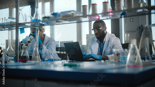 Bright Medical Science Laboratory with Brilliant Team of Research Scientists Working. Microbiologist Looks Under Microscope Talks to Biochemist Using Laptop. High-Tech Equipment.