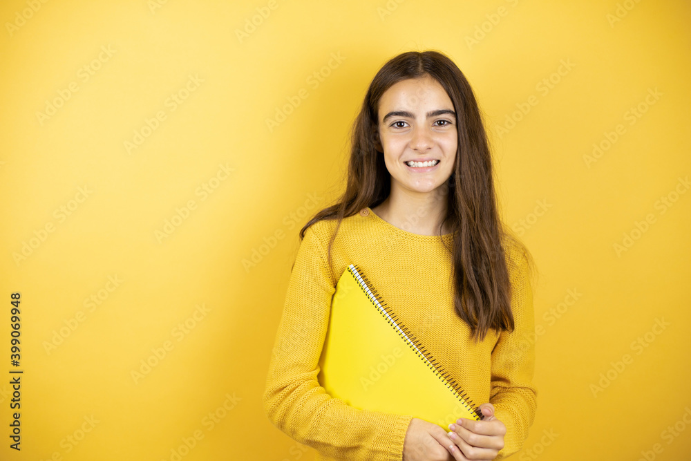 Pretty girl wearing a yellow sweater standing over isolated yellow background holding a notebook Pretty girl wearing a yellow sweater standing over isolated yellow background holding a notebook