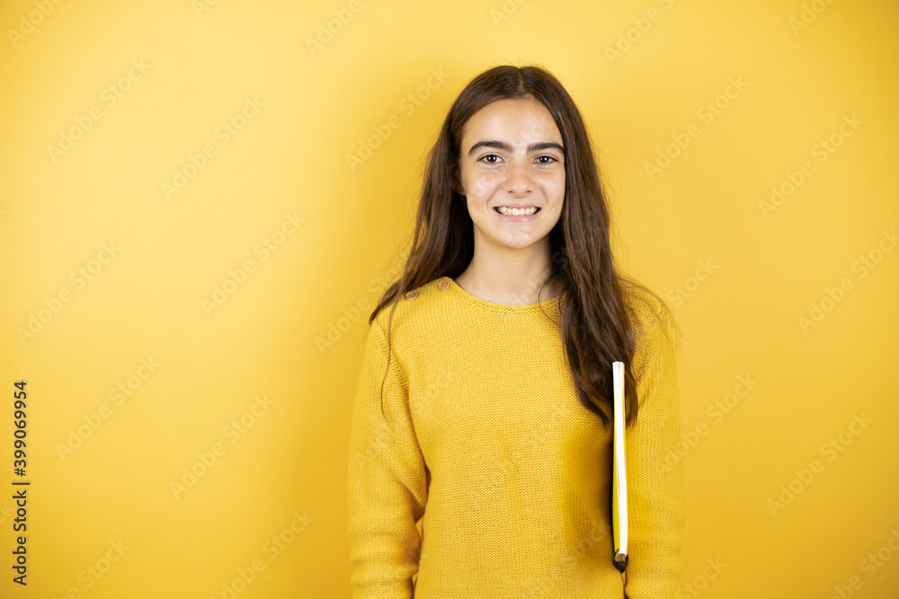 Pretty girl wearing a yellow sweater standing over isolated yellow background holding a notebook Pretty girl wearing a yellow sweater standing over isolated yellow background holding a notebook
