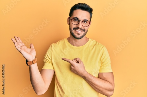 Young hispanic man wearing casual clothes and glasses amazed and smiling to the camera while presenting with hand and pointing with finger.