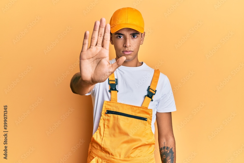 Young handsome african american man wearing handyman uniform over yellow background doing stop sing with palm of the hand. warning expression with negative and serious gesture on the face. Young handsome african american man wearing handyman uniform over yellow background doing stop sing with palm of the hand. warning expression with negative and serious gesture on the face.