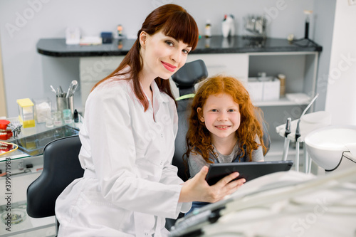 Medicine, pediatric dentistry and oral care concept. Female smiling dentist showing tablet pc computer to happy kid patient, curly red haired school girl at modern dental clinic