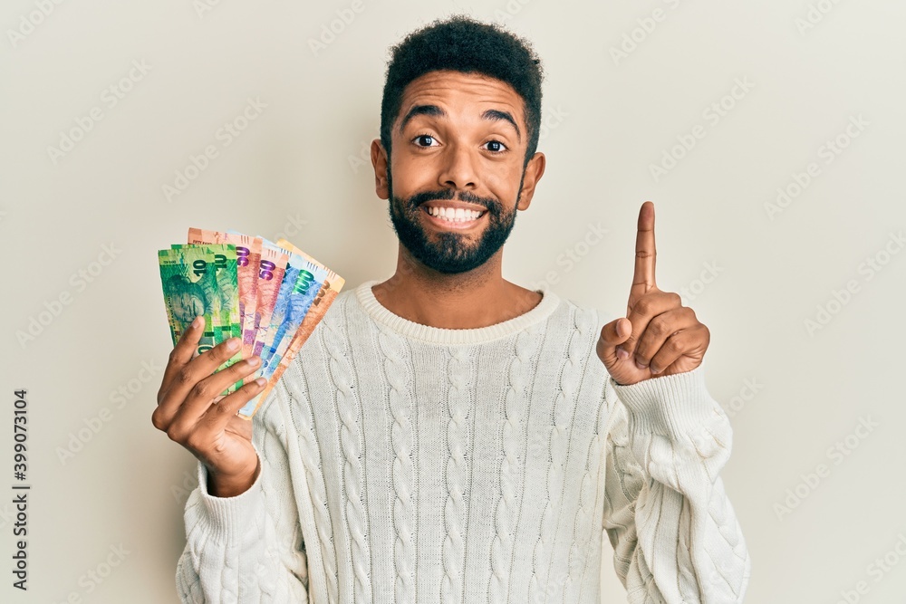 Handsome hispanic man with beard holding south african rand banknotes smiling with an idea or question pointing finger with happy face, number one Handsome hispanic man with beard holding south african rand banknotes smiling with an idea or question pointing finger with happy face, number one