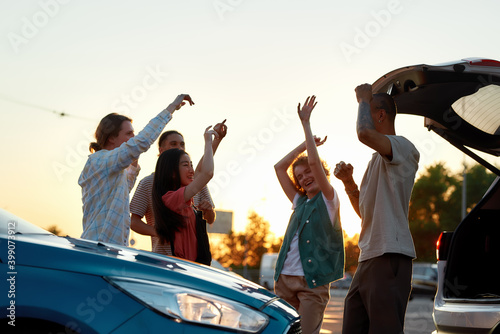 Five young casually dressed friends having a party dancing to music together outside on a parking site near their cars