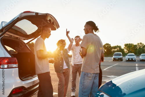 Five young commonly dressed friends outside on a parking site speaking to each other laughing and dancing near their cars with an opened trunk