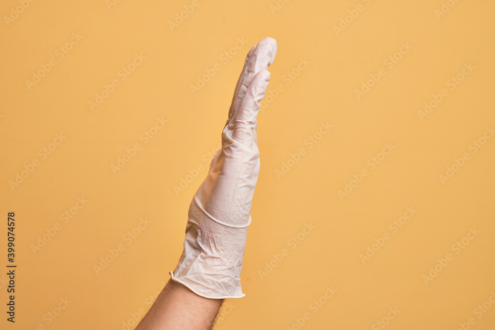 Hand of caucasian young man with medical glove over isolated yellow background showing side of stretched hand, pushing and doing stop gesture Hand of caucasian young man with medical glove over isolated yellow background showing side of stretched hand, pushing and doing stop gesture