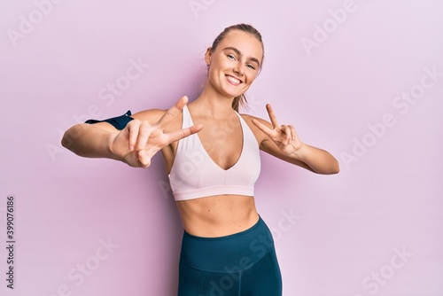Beautiful blonde woman wearing sportswear and arm band smiling looking to the camera showing fingers doing victory sign. number two.