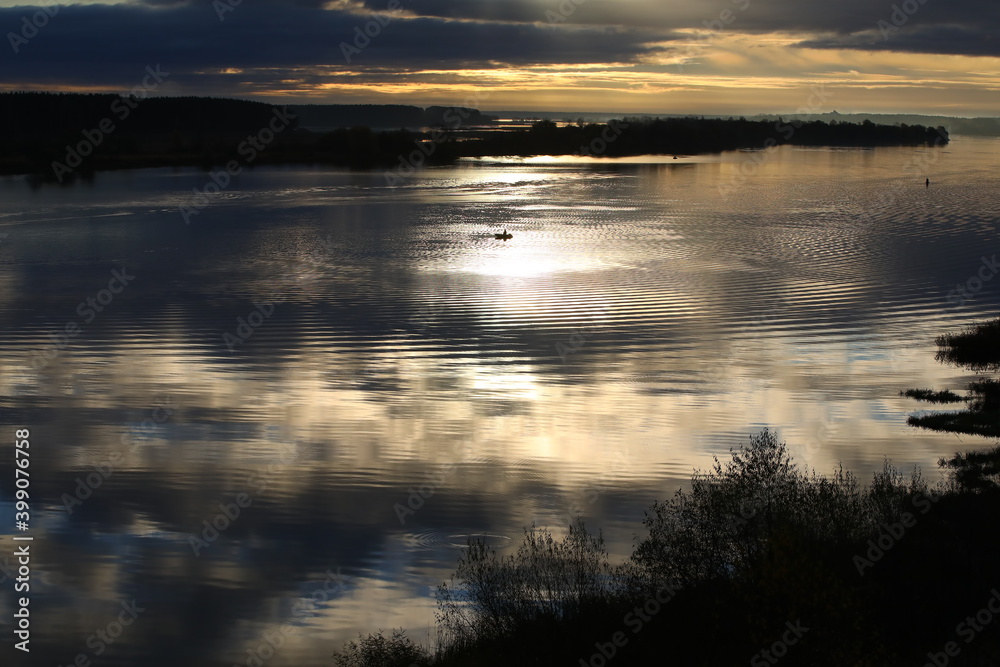 Fototapeta premium Night river.The black sky with Golden stripes is reflected in the water with ripples on the surface.In the center of the water area is a distant silhouette of a fisherman in a boat in the moonlight