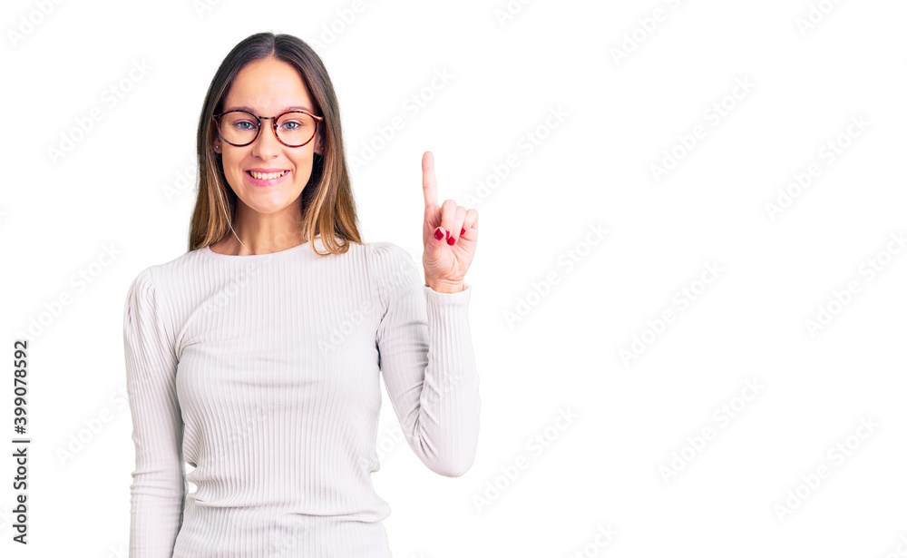 Obraz premium Beautiful brunette young woman wearing casual white sweater and glasses showing and pointing up with finger number one while smiling confident and happy.