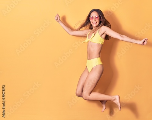 Young beautiful girl wearing bikini and sunglasses smiling happy. Jumping with smile on face celebrating with fists up over isolated yellow background
