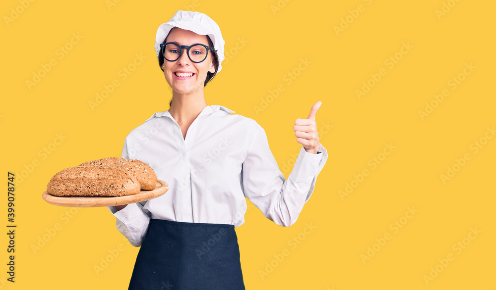 Beautiful brunette young woman wearing baker uniform holding homemade bread smiling happy and positive, thumb up doing excellent and approval sign Beautiful brunette young woman wearing baker uniform holding homemade bread smiling happy and positive, thumb up doing excellent and approval sign