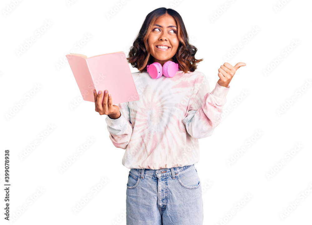 Young beautiful mixed race woman reading a book wearing headphones pointing thumb up to the side smiling happy with open mouth Young beautiful mixed race woman reading a book wearing headphones pointing thumb up to the side smiling happy with open mouth