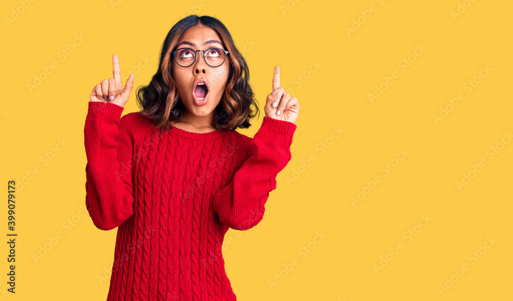 Young beautiful mixed race woman wearing red sweater and glasses amazed and surprised looking up and pointing with fingers and raised arms. Young beautiful mixed race woman wearing red sweater and glasses amazed and surprised looking up and pointing with fingers and raised arms.