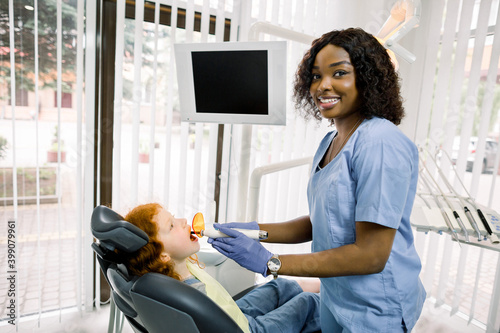 Medicine  stomatology and teeth care concept. Young smiling African woman dentist with dental curing UV light lamp  looking at camera  finishing tooth repair for her little patient  cute pretty girl