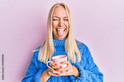 Young blonde girl wearing winter sweater and drinking a cup of hot coffee sticking tongue out happy with funny expression.