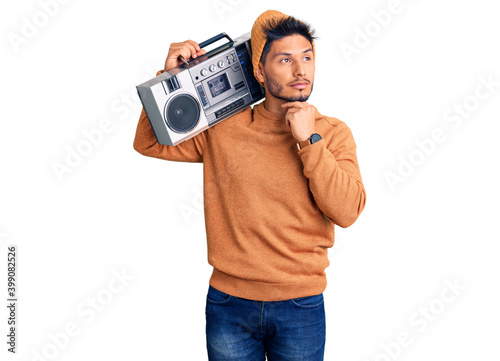 Handsome latin american young man holding boombox, listening to music with hand on chin thinking about question, pensive expression. smiling with thoughtful face. doubt concept.