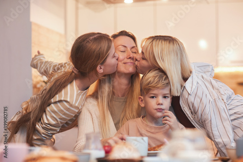 Portrait of happy mother enjoying kisses from three children while sitting at breakfast table in kitchen