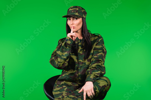 Young beautiful woman in military uniform posing while sitting on a chair, gesturing shh with index finger. Gender stereotypes.