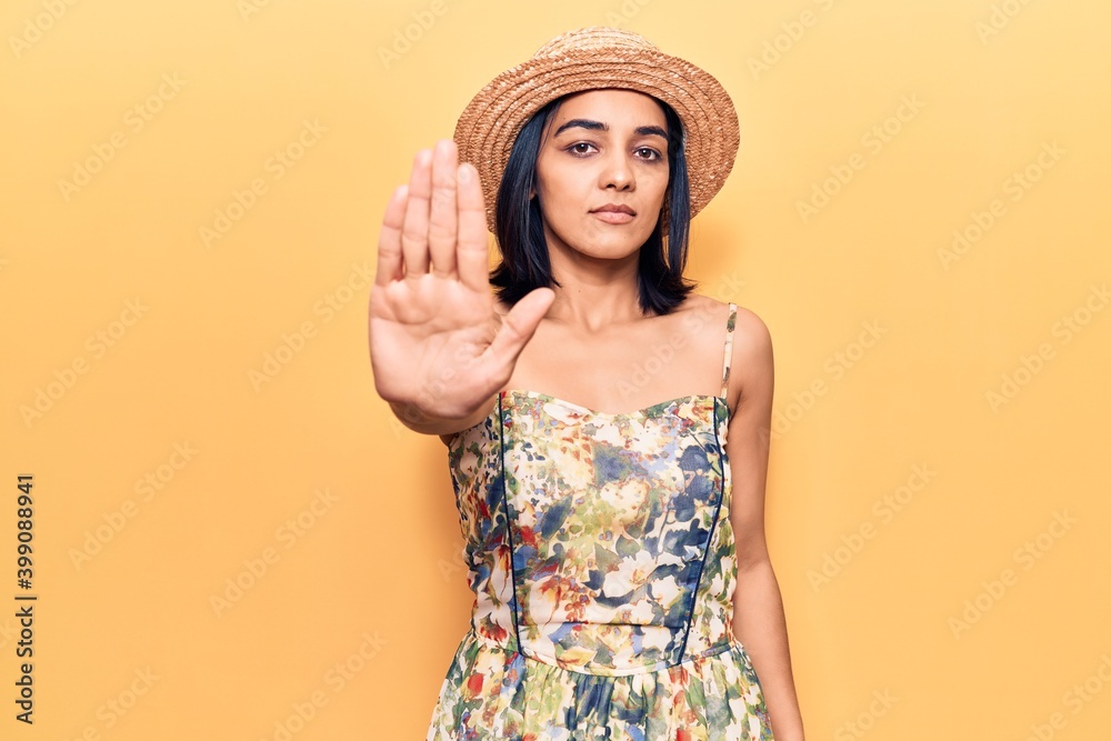 Young beautiful latin woman wearing summer hat doing stop sing with palm of the hand. warning expression with negative and serious gesture on the face. Young beautiful latin woman wearing summer hat doing stop sing with palm of the hand. warning expression with negative and serious gesture on the face.