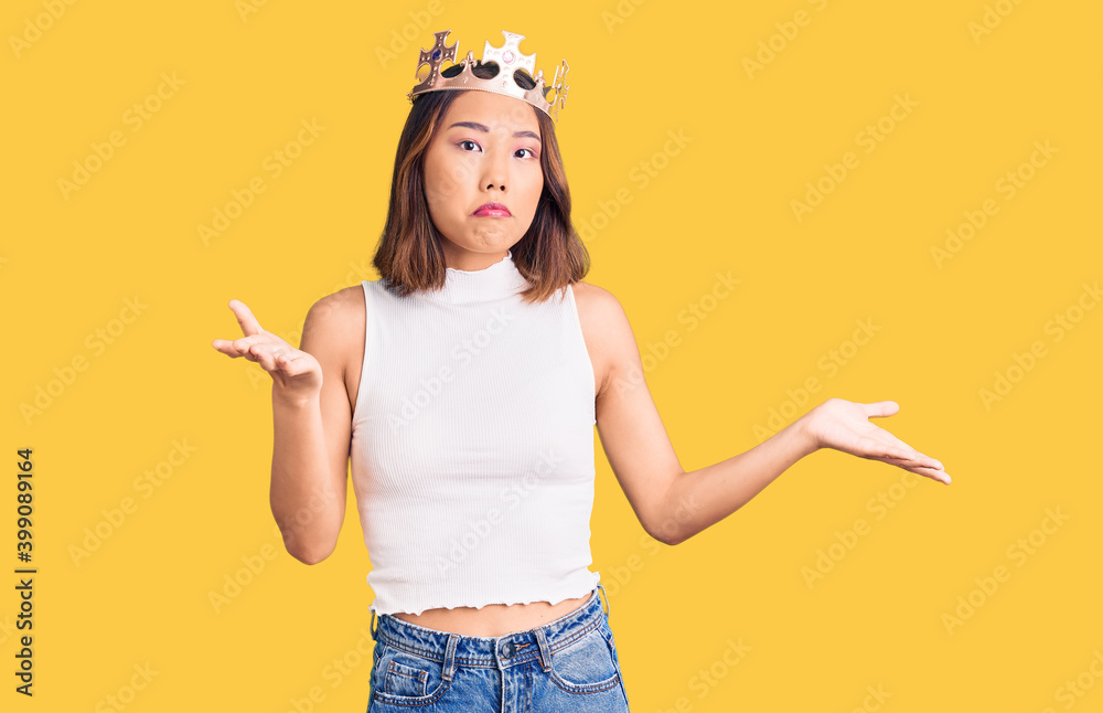 Young beautiful chinese girl wearing princess crown clueless and confused expression with arms and hands raised. doubt concept.