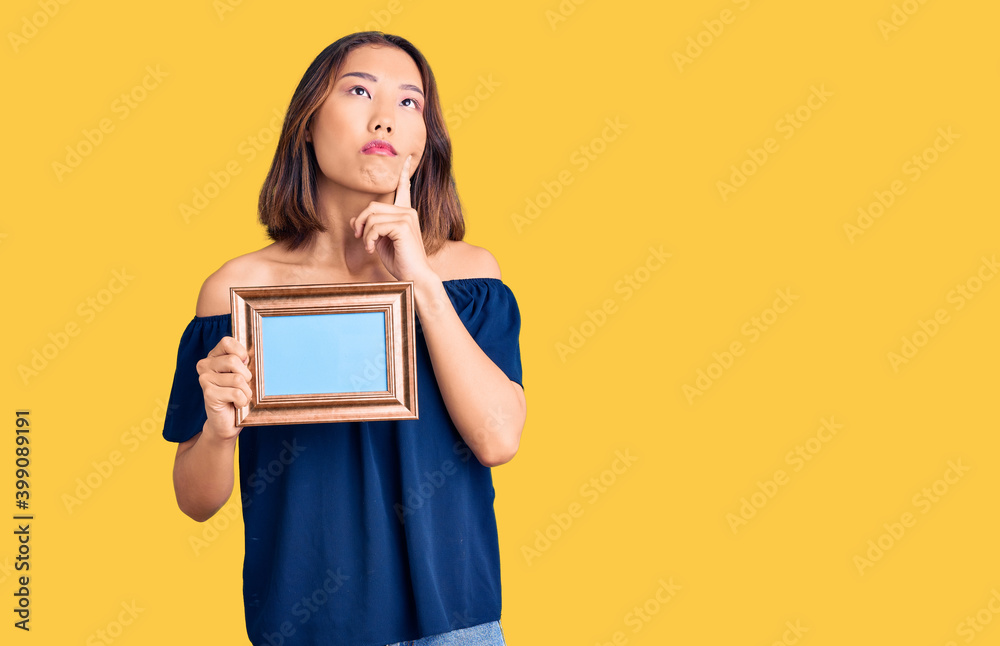 Young beautiful chinese girl holding empty frame serious face thinking about question with hand on chin, thoughtful about confusing idea Young beautiful chinese girl holding empty frame serious face thinking about question with hand on chin, thoughtful about confusing idea