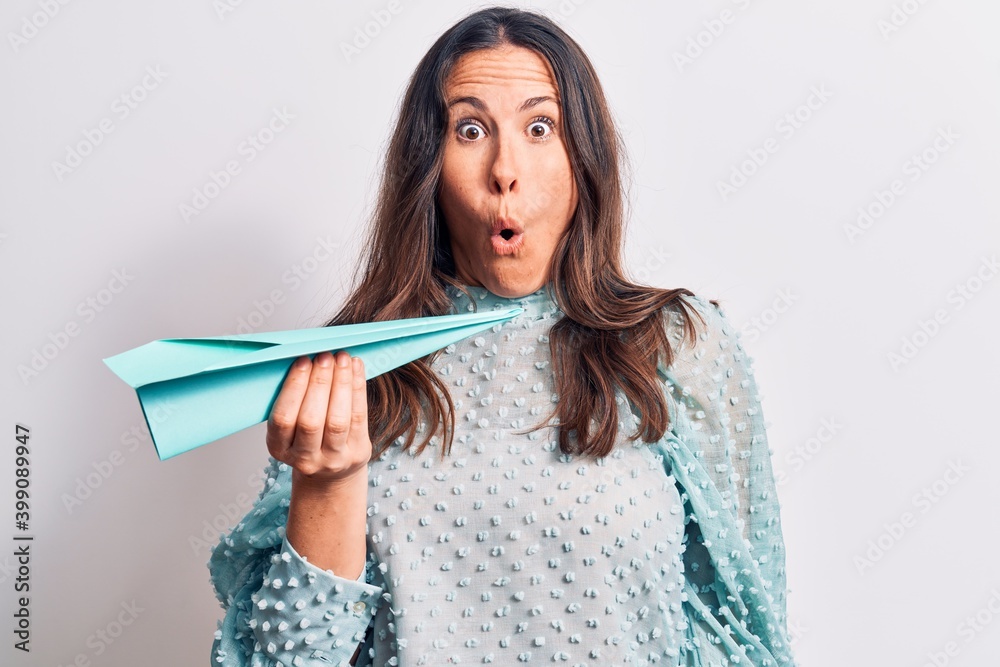 Young beautiful brunette woman holding paper airplane over isolated white background scared and amazed with open mouth for surprise, disbelief face Young beautiful brunette woman holding paper airplane over isolated white background scared and amazed with open mouth for surprise, disbelief face