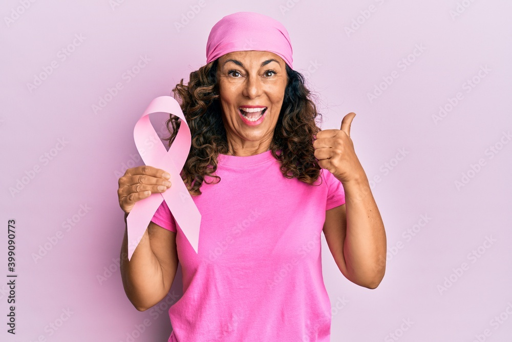 Middle age hispanic woman holding pink cancer ribbon pointing thumb up to the side smiling happy with open mouth Middle age hispanic woman holding pink cancer ribbon pointing thumb up to the side smiling happy with open mouth