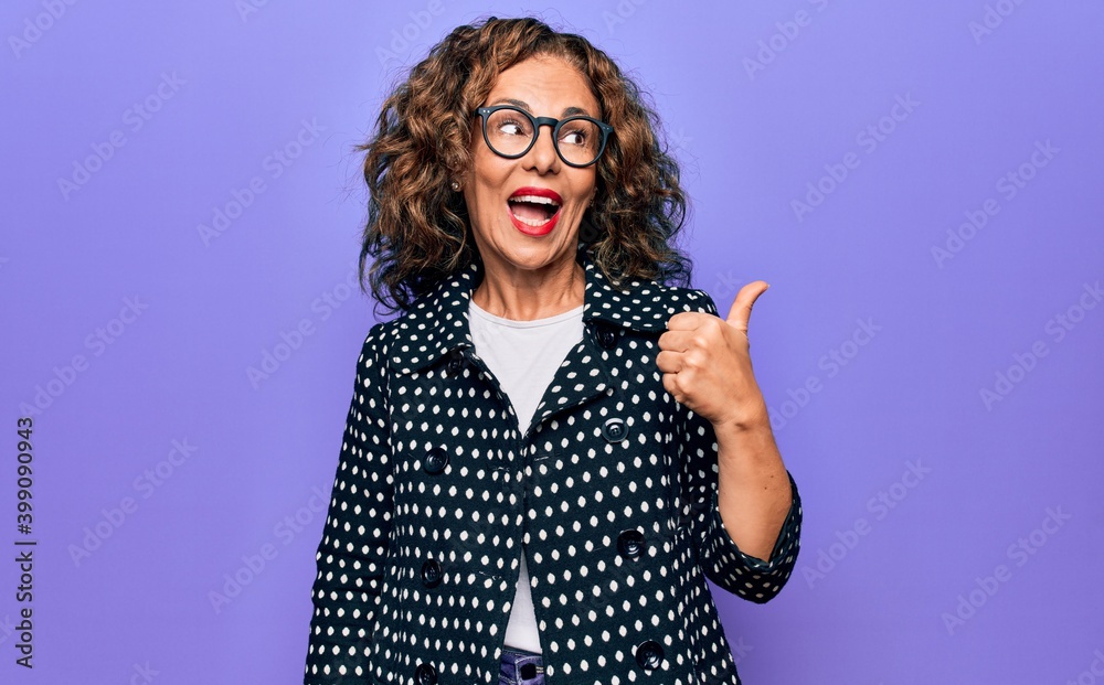 Middle age beautiful woman wearing casual coat and glasses over isolated purple background pointing thumb up to the side smiling happy with open mouth Middle age beautiful woman wearing casual coat and glasses over isolated purple background pointing thumb up to the side smiling happy with open mouth
