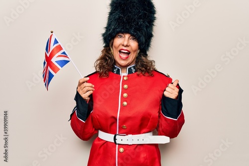 Middle age beautiful wales guard woman wearing traditional uniform holding united kingdom flag pointing thumb up to the side smiling happy with open mouth