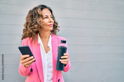 Middle age hispanic businesswoman with serious expression using smartphone and drinking bottle of water at the city.