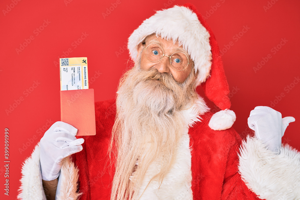 Old senior man with grey hair and long beard wearing santa claus costume holding boarding pass pointing thumb up to the side smiling happy with open mouth