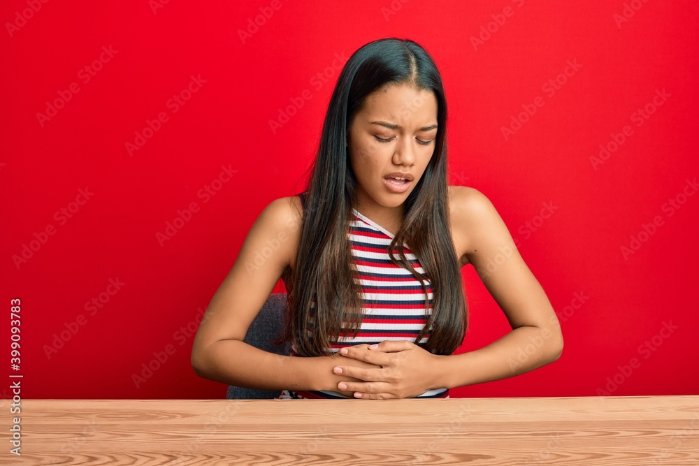 Beautiful hispanic woman wearing casual clothes sitting on the table with hand on stomach because nausea, painful disease feeling unwell. ache concept.