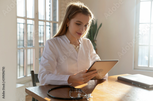 Young Caucasian female doctor in white medical uniform sit at desk in hospital consulting client online on tablet. Woman GP or nurse use pad device have video call on gadget with clinic patient. © fizkes