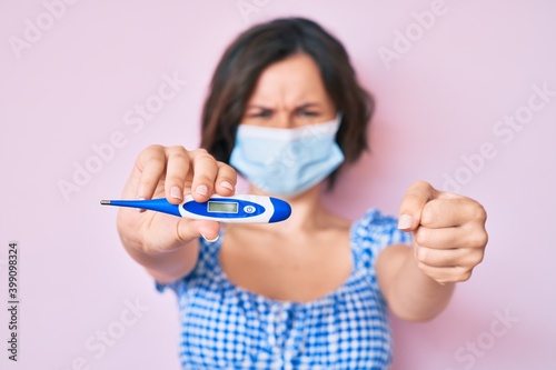 Young beautiful woman wearing medical mask holding holding thermometer annoyed and frustrated shouting with anger, yelling crazy with anger and hand raised